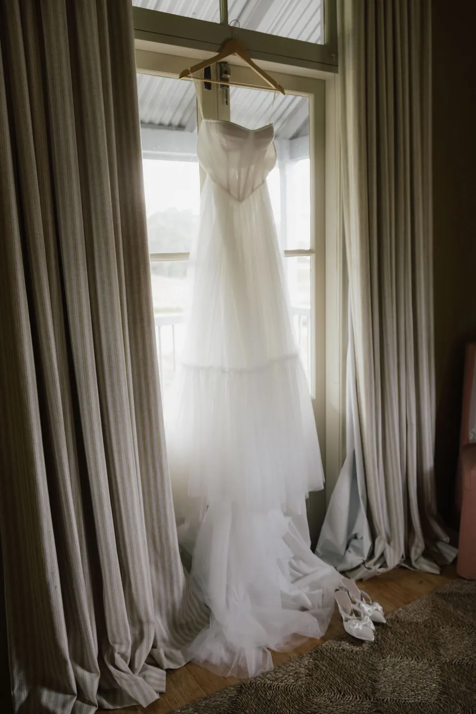 Bridget's wedding dress hanging in the window of a country cottage. At Cudgera Creek in northern New South Wales, for an Elopement Collective elopement. Photograph by Bulb Creative.