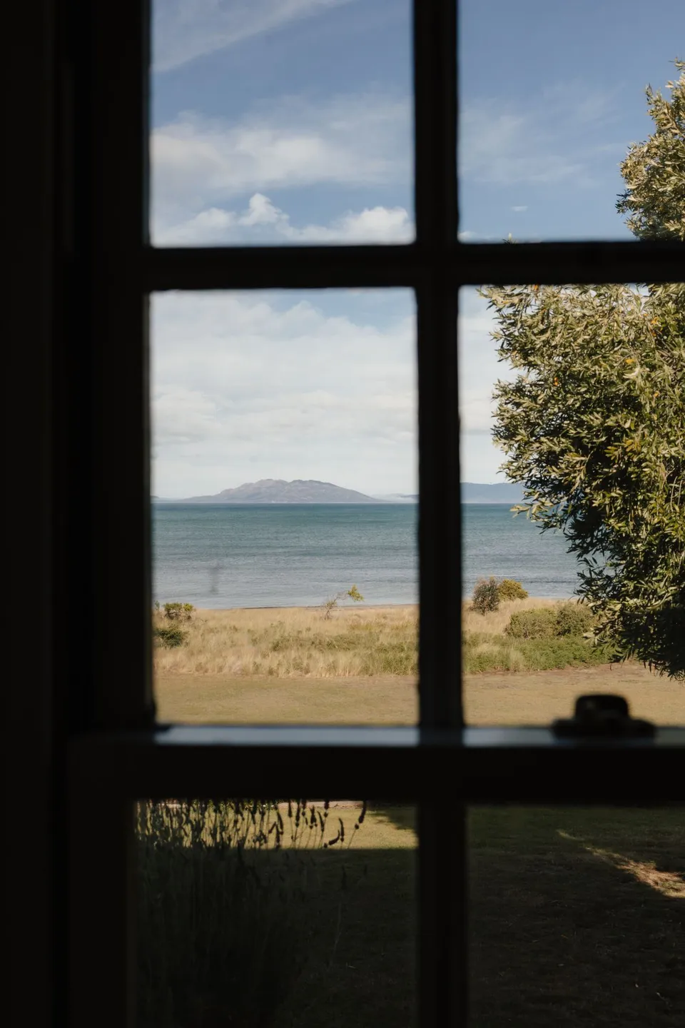 Great Oyster Bay and distant hills seen through a dark timber-framed cottage window. At Piermont Retreat on Tasmania’s east coast, for an Elopement Collective elopement. Photograph by Bulb Creative.
