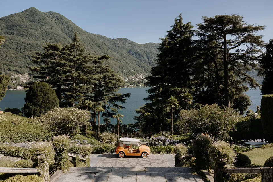 An orange open-top car on a cobbled driveway among terraced gardens and tall trees overlooking the lake, at Passalacqua in Moltrasio, Lake Como, Italy, for an Elopement Collective elopement. Photo by George Bowden.