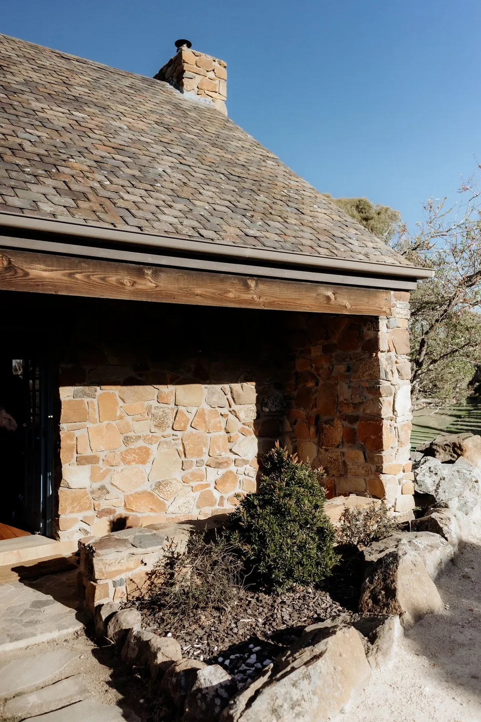 A sunlit stone cottage with a slate roof, chimney and garden beneath a clear blue sky. At Piermont Retreat on Tasmania’s east coast, for an Elopement Collective elopement. Photograph by Omar of By The Wilde.