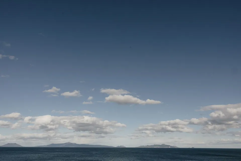 Blue water in Great Oyster Bay beneath a broad sky with scattered clouds and distant coastal hills, at Thalia Haven on the East Coast of Tasmania, for an Elopement Collective elopement. Photograph by Bulb Creative.