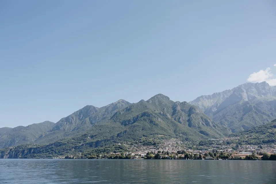 Blue water, wooded mountain slopes and a lakeside village seen from across Lake Como, at Lake Como in Lombardy, Italy, for an Elopement Collective elopement. Photograph by George Bowden.