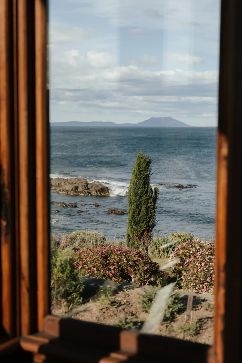A view through a timber-framed window towards a cypress tree, the rocky shoreline and Great Oyster Bay, at Thalia Haven on Tasmania’s east coast, for an Elopement Collective elopement. Photograph by Bulb Creative.