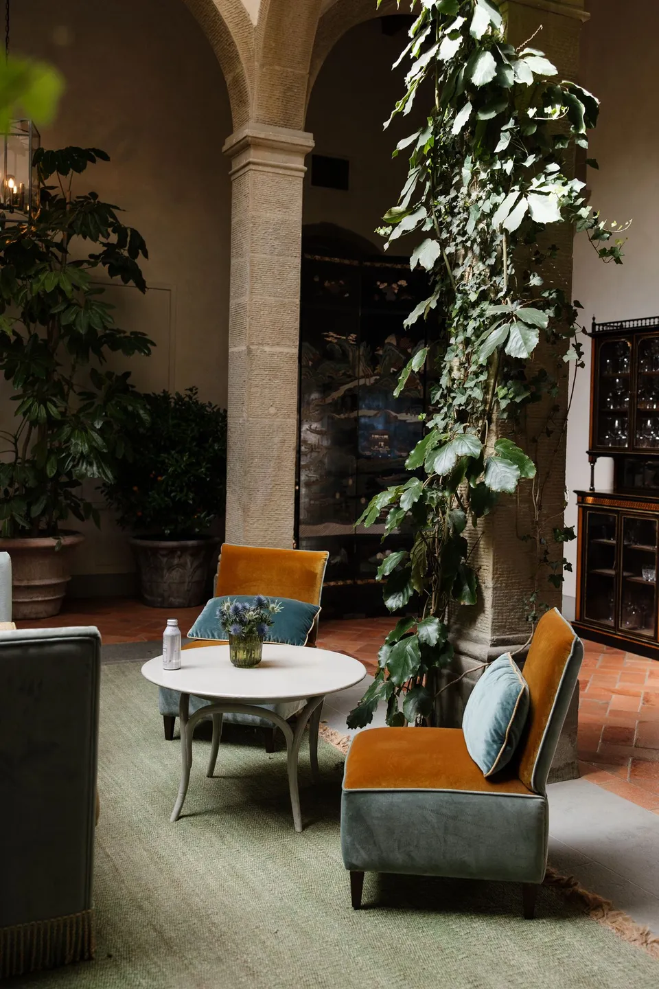 A stone arch and column beside a lounge chair, small table and trailing greenery in the villa cloister, at Villa San Michele in Fiesole above Florence, Tuscany, Italy, for an Elopement Collective elopement. Digital photograph by Khara from House of Lucie.