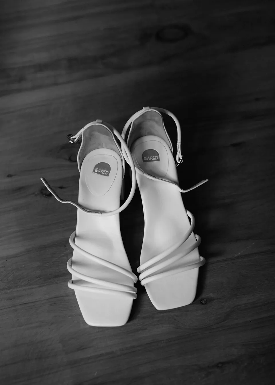 Black-and-white detail of Elyse's white strappy wedding shoes on the timber floor. At Fig Tree Villa in Ewingsdale, Byron Bay, for an Elopement Collective elopement. Photograph by Bulb Creative.