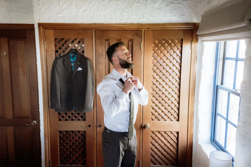 Russell adjusting his tie beside the cottage window, with his jacket hanging behind him. At Piermont Retreat on Tasmania’s east coast, for an Elopement Collective elopement. Photograph by Omar of By The Wilde.