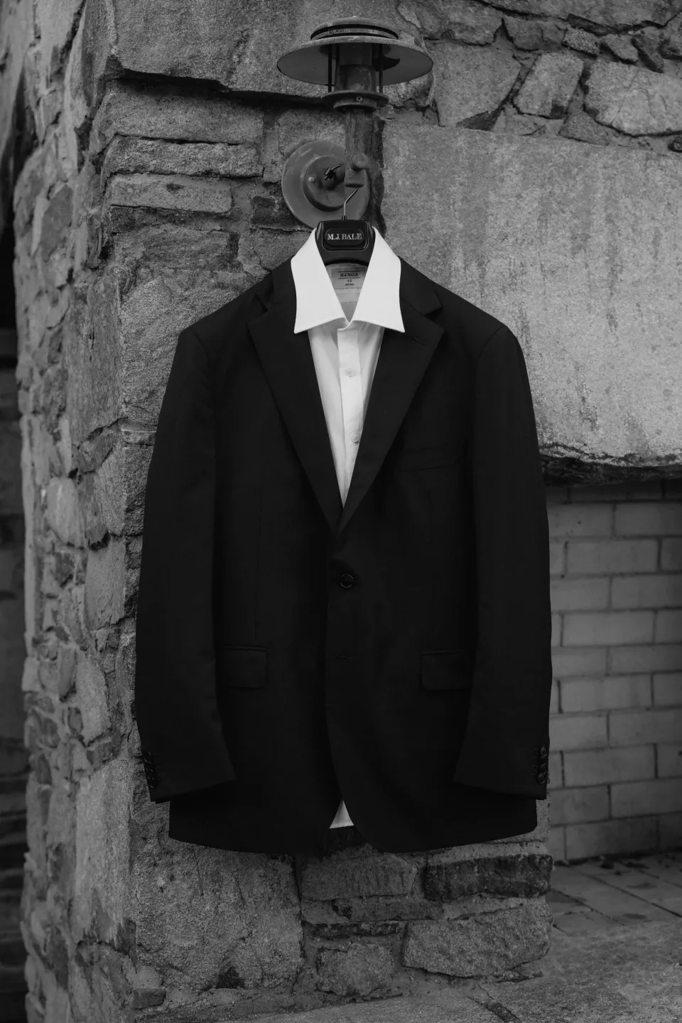 Black and white photograph of Anthony’s dark suit jacket and white shirt hanging against a stone wall, at Thalia Haven on Tasmania’s east coast, for an Elopement Collective elopement. Photograph by Bulb Creative.