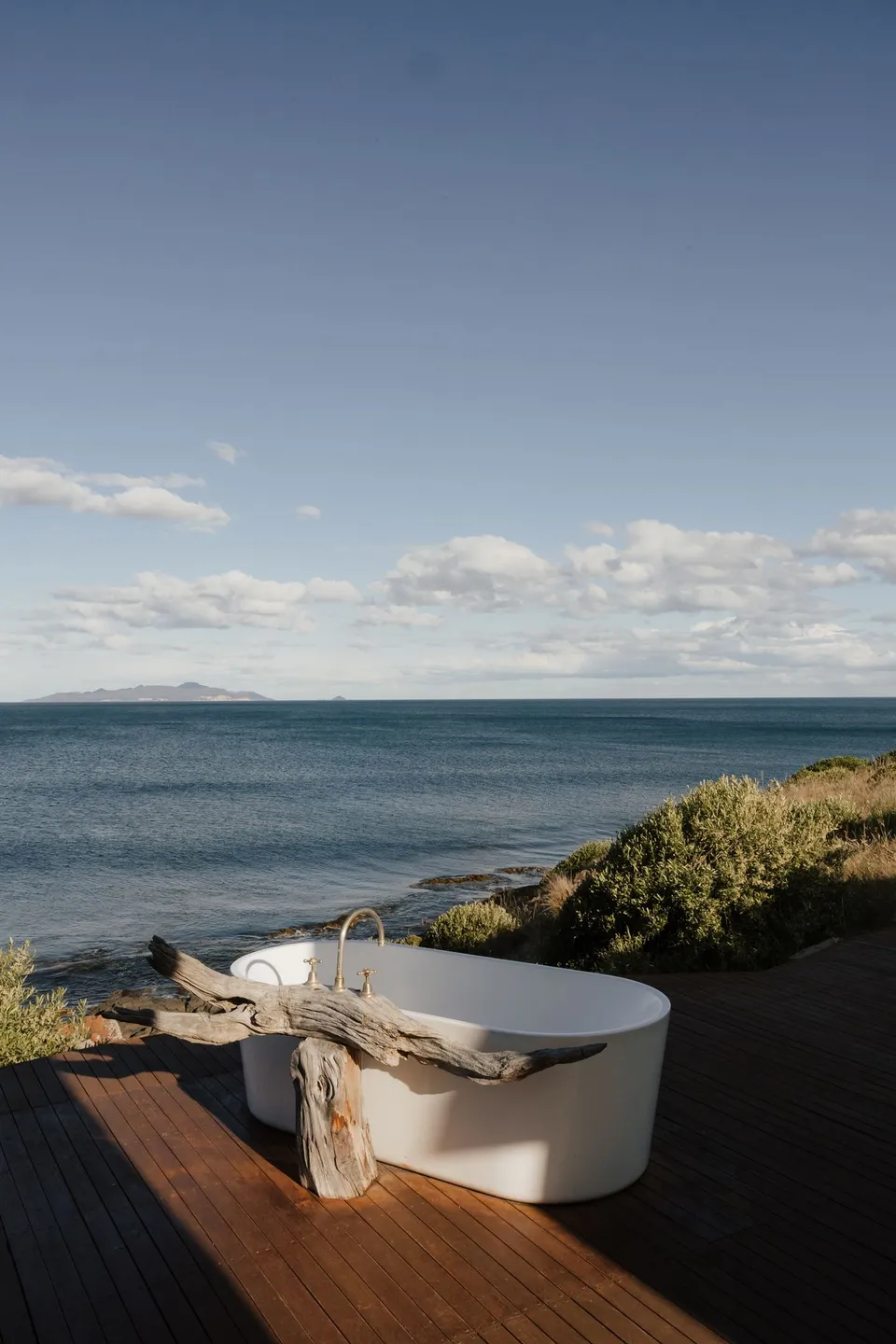 Thalia Haven’s white outdoor bathtub and driftwood stand on a timber deck overlooking blue water, at Thalia Haven on the East Coast of Tasmania, for an Elopement Collective elopement. Photograph by Bulb Creative.