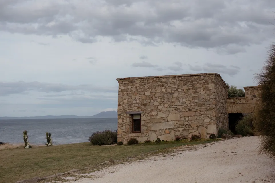 A stone cottage beside the bay beneath an overcast sky, at Thalia Haven on the East Coast of Tasmania, Australia, for an Elopement Collective elopement. Photograph by Joey Willis.