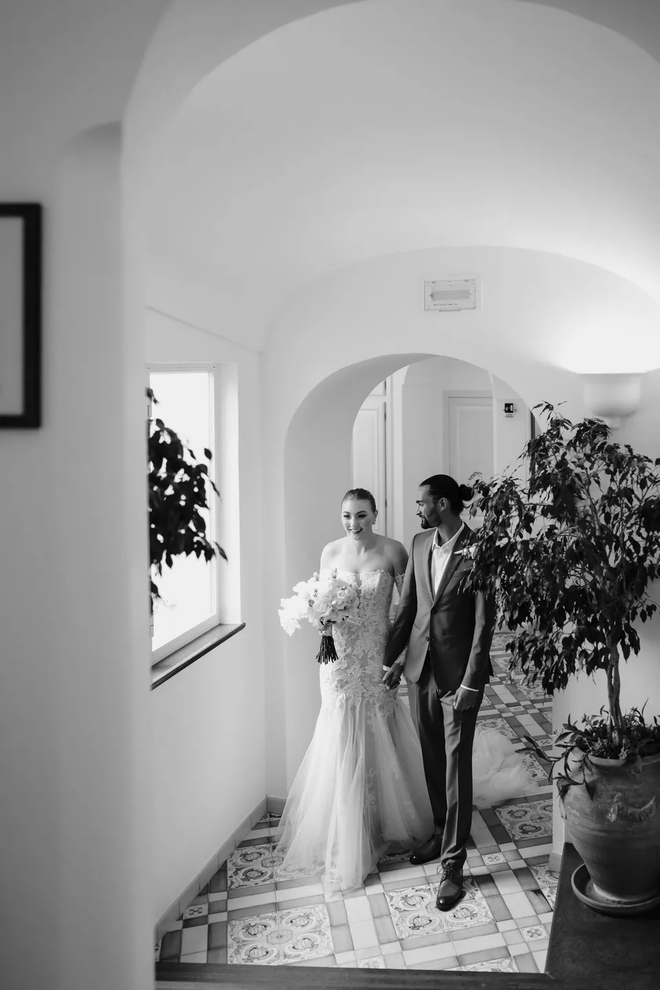 Black and white portrait of Skyler and Zach standing together beneath an arched ceiling in the hotel hallway, at Hotel Buca di Bacco in Positano, Amalfi Coast, Italy, for an Elopement Collective elopement. Photograph by Jason Corroto.