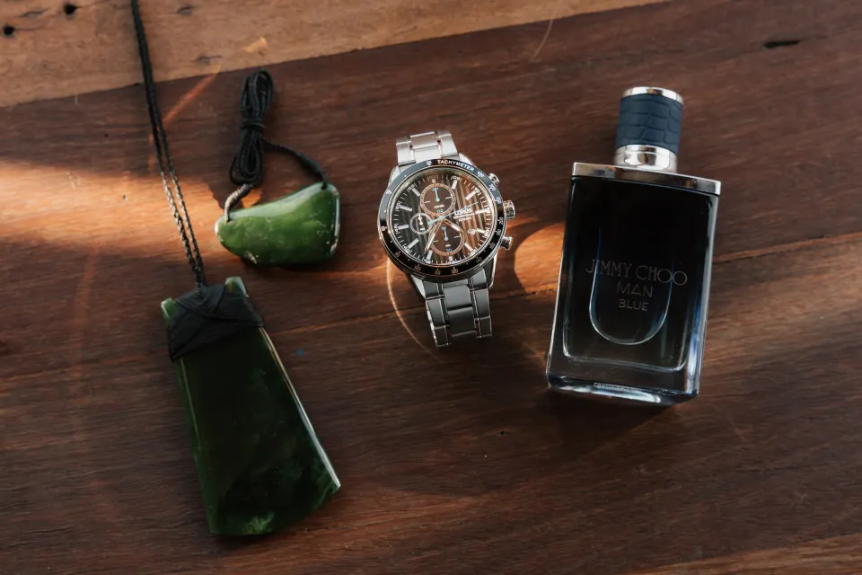 Two green pendants, a silver watch and a dark fragrance bottle arranged on sunlit timber, at Thalia Haven on Tasmania’s east coast, for an Elopement Collective elopement. Photograph by Bulb Creative.