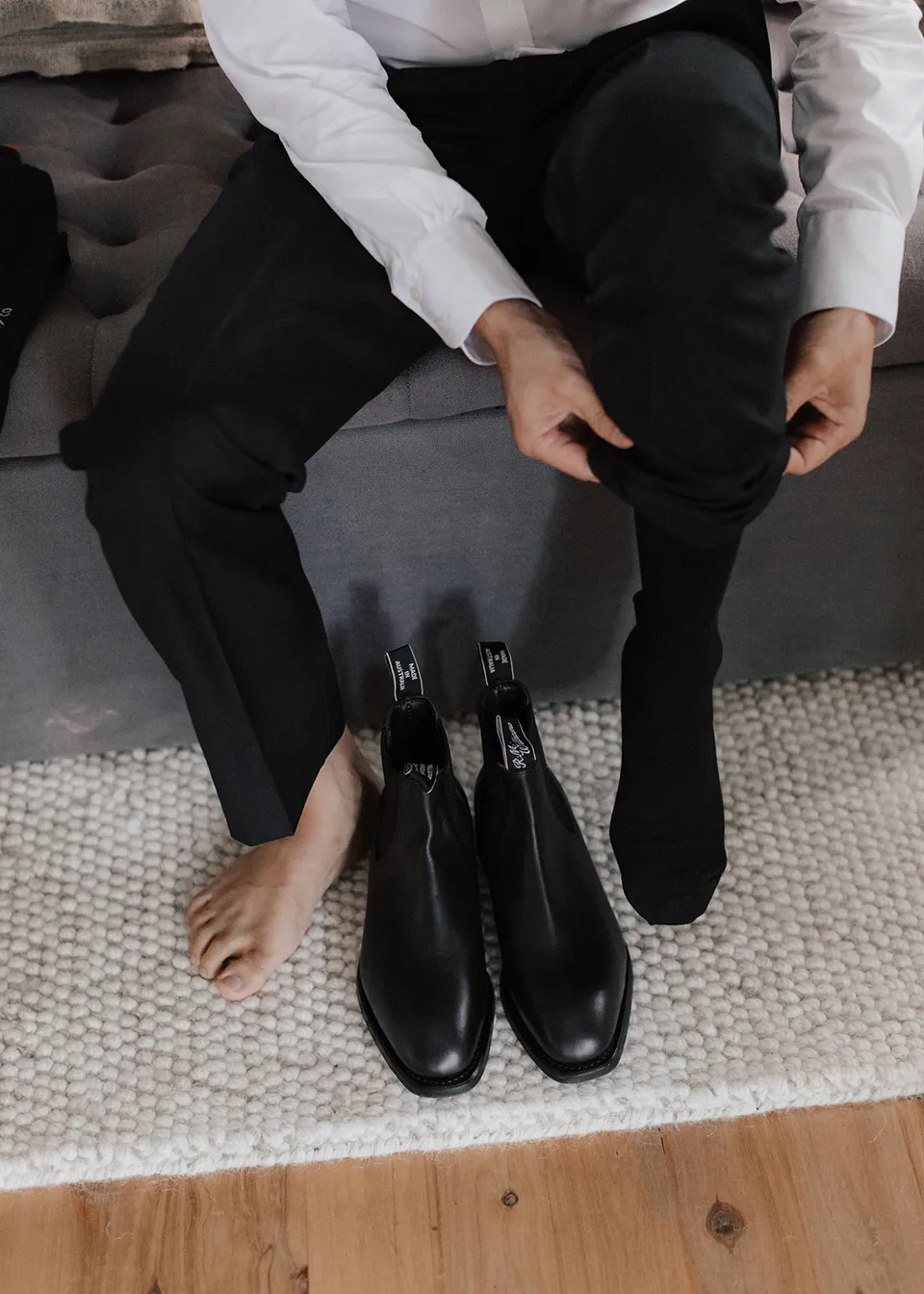 Jonathan adjusting his trouser leg with polished black boots on the floor. At Fig Tree Villa in Ewingsdale, Byron Bay, for an Elopement Collective elopement. Photograph by Bulb Creative.