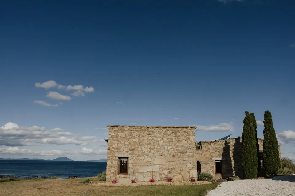 A stone building and tall cypress trees above golden grassland, with Great Oyster Bay behind them, at Thalia Haven on the East Coast of Tasmania, for an Elopement Collective elopement. Photograph by Bulb Creative.