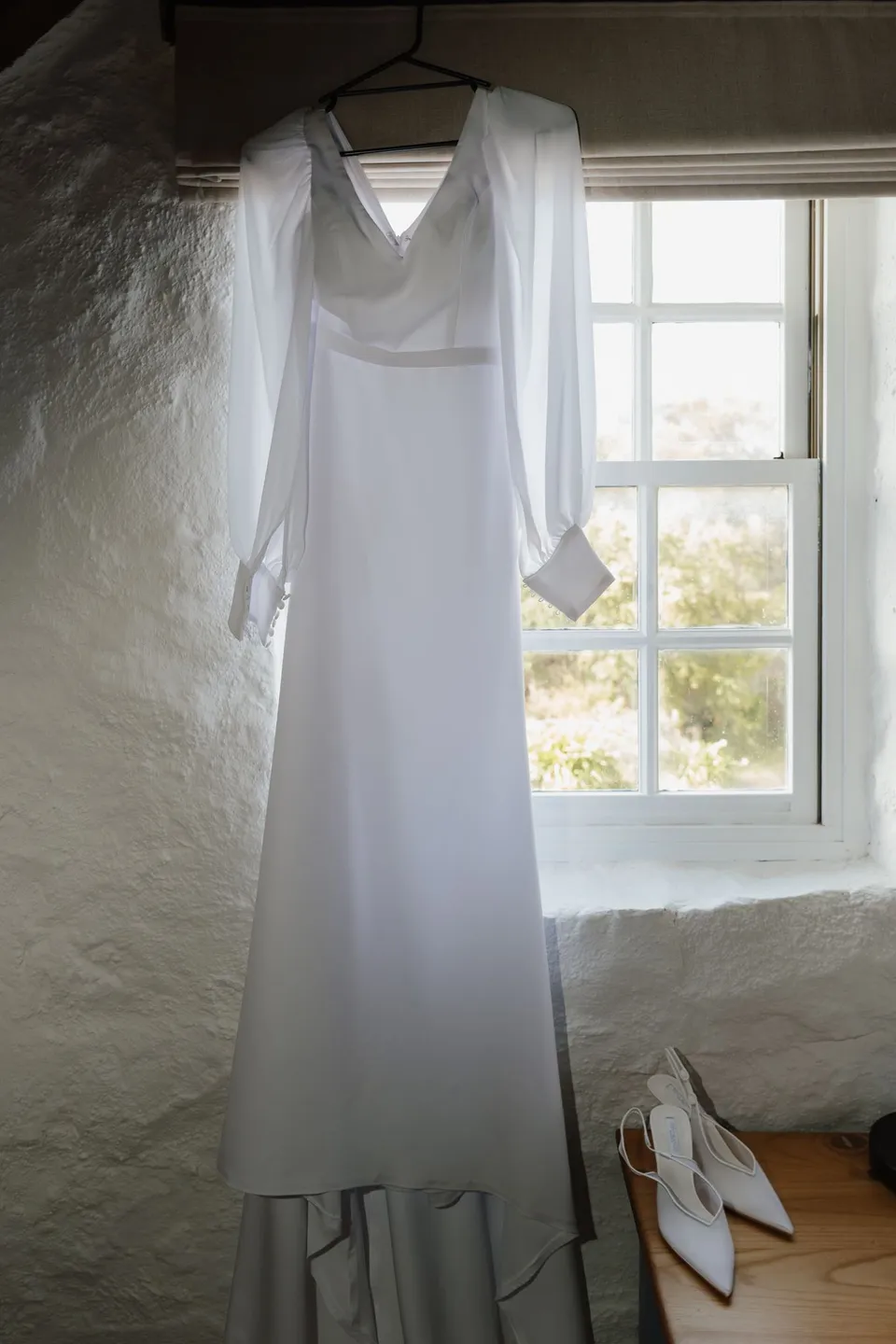Renee’s long-sleeved wedding dress hanging beside a sunlit cottage window, with her shoes below. At Piermont Retreat on Tasmania’s east coast, for an Elopement Collective elopement. Photograph by Bulb Creative.