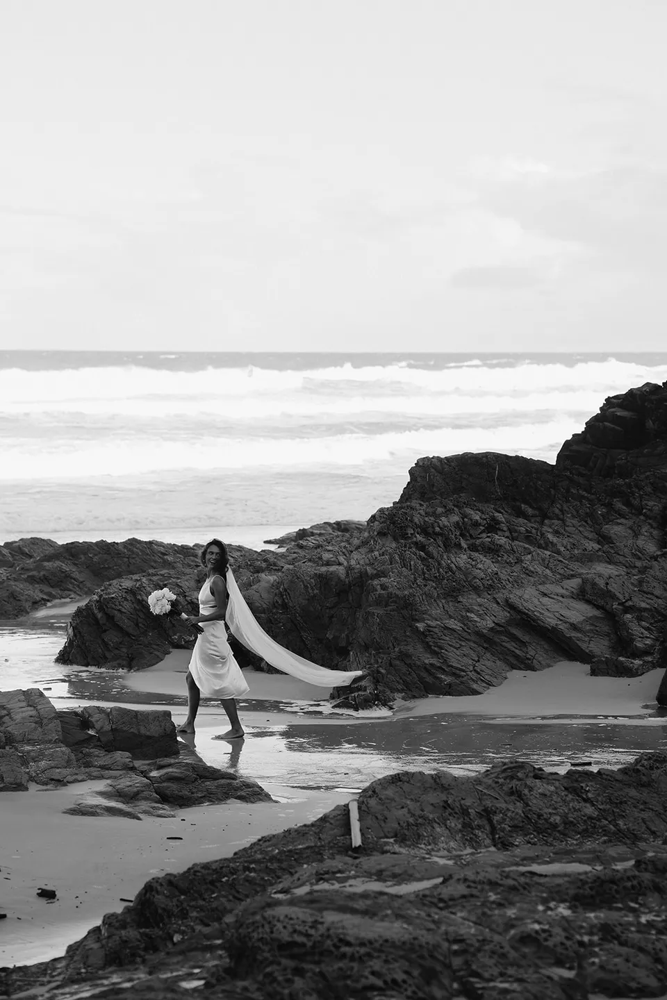 Black-and-white portrait of Rachel standing among the coastal rocks with her veil trailing behind her. At Whites Beach, Broken Head, near Byron Bay, for an Elopement Collective elopement. Photograph by Jason Lucas.