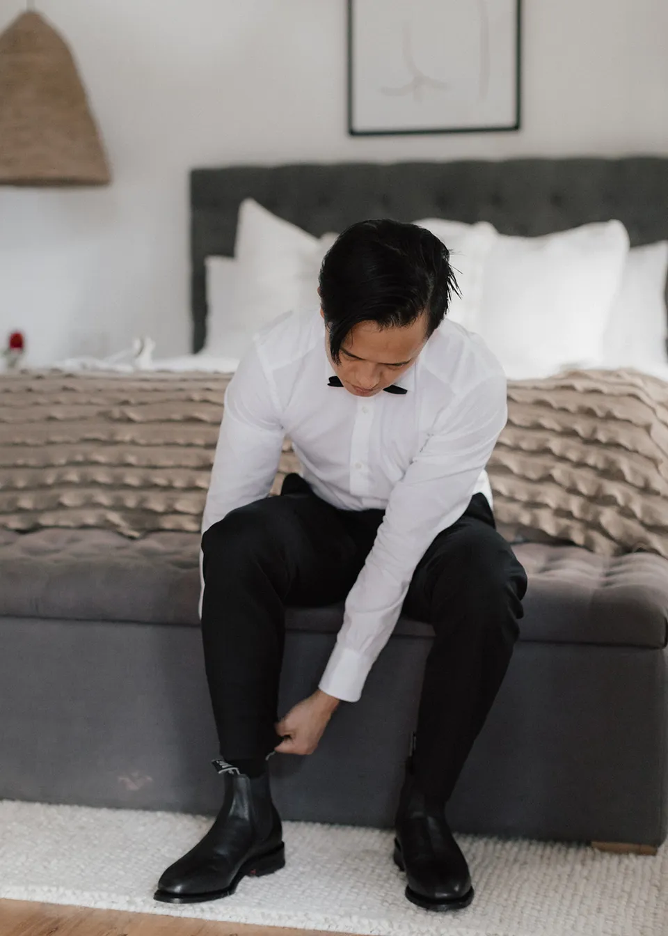 Jonathan fastening his black boot beside the bed. At Fig Tree Villa in Ewingsdale, Byron Bay, for an Elopement Collective elopement. Photograph by Bulb Creative.