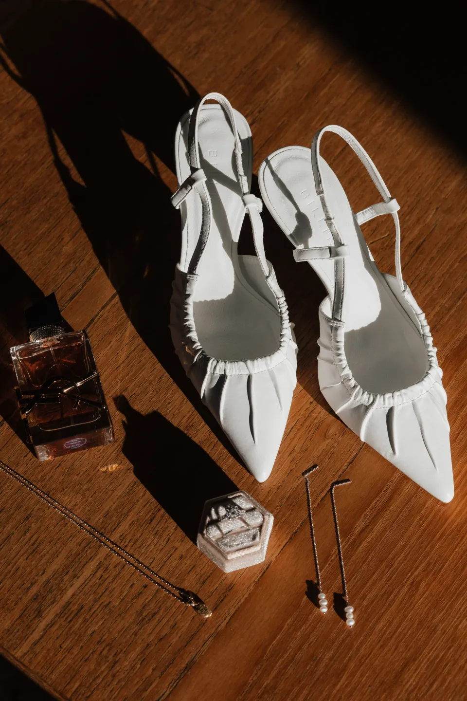 Dani’s white pointed slingback shoes, jewellery and fragrance arranged on a wooden floor, at Thalia Haven on Tasmania’s east coast, for an Elopement Collective elopement. Photograph by Bulb Creative.