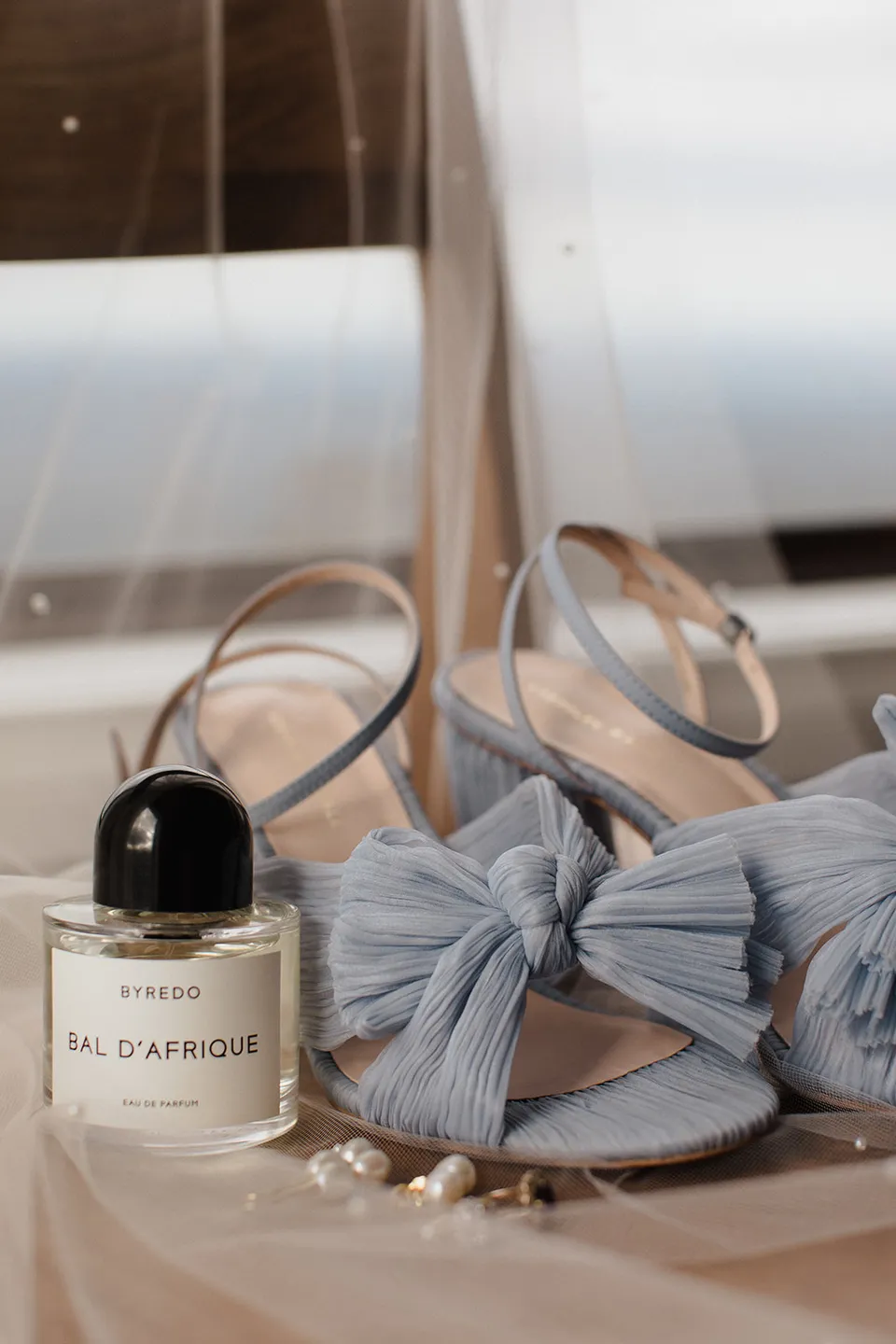 Meg’s pale blue bow-front shoes, fragrance and jewellery arranged beside a window. At The Keep on Tasmania’s east coast, for an Elopement Collective elopement. Photograph by Jenna Agius.