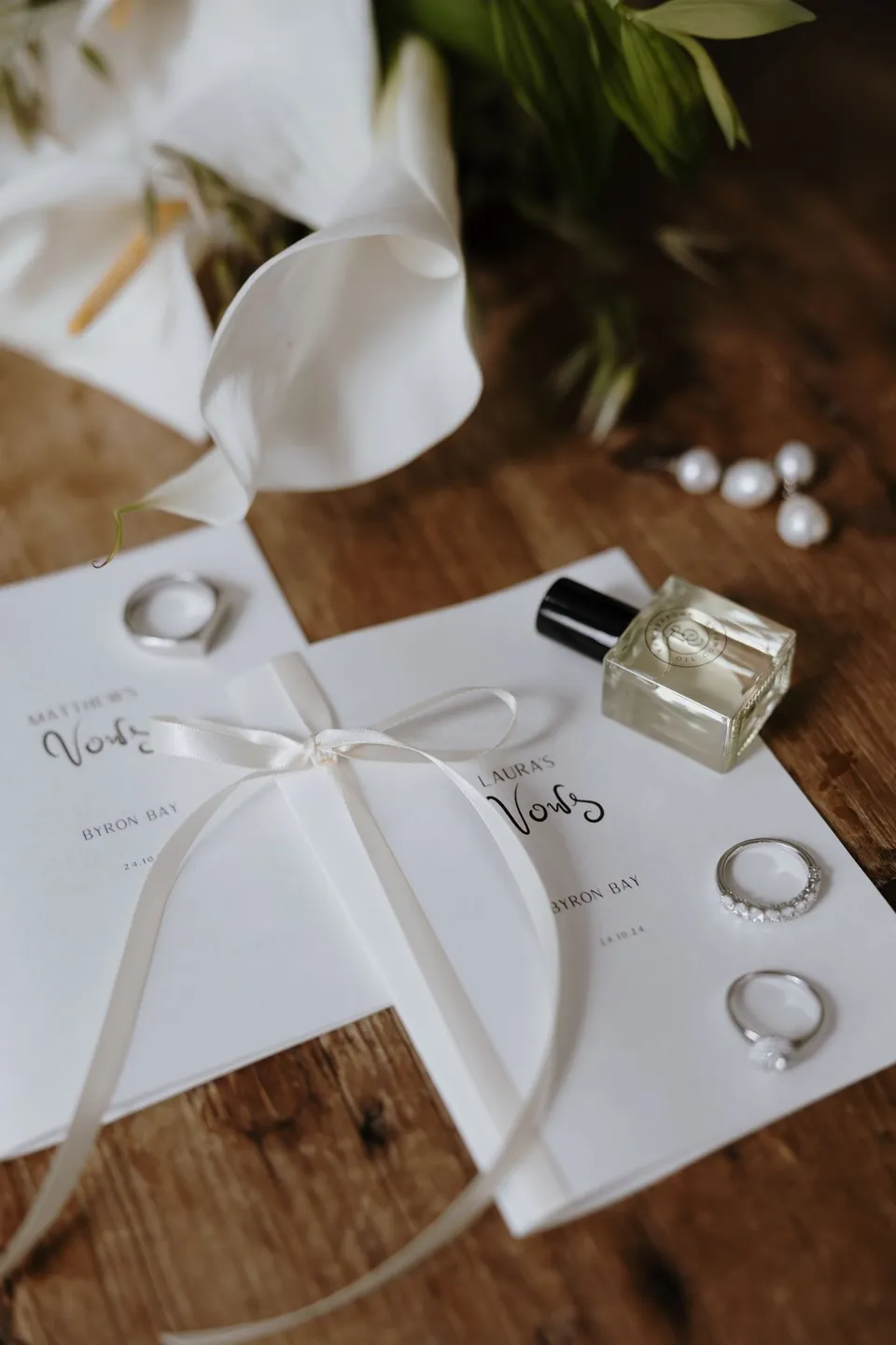 Wedding rings, perfume and vow booklets tied with a white ribbon beside pearl earrings and white flowers by The French Petal on a timber table. At Fig Tree Villa in Ewingsdale, Byron Bay, for an Elopement Collective elopement. Photograph by George Bowden.