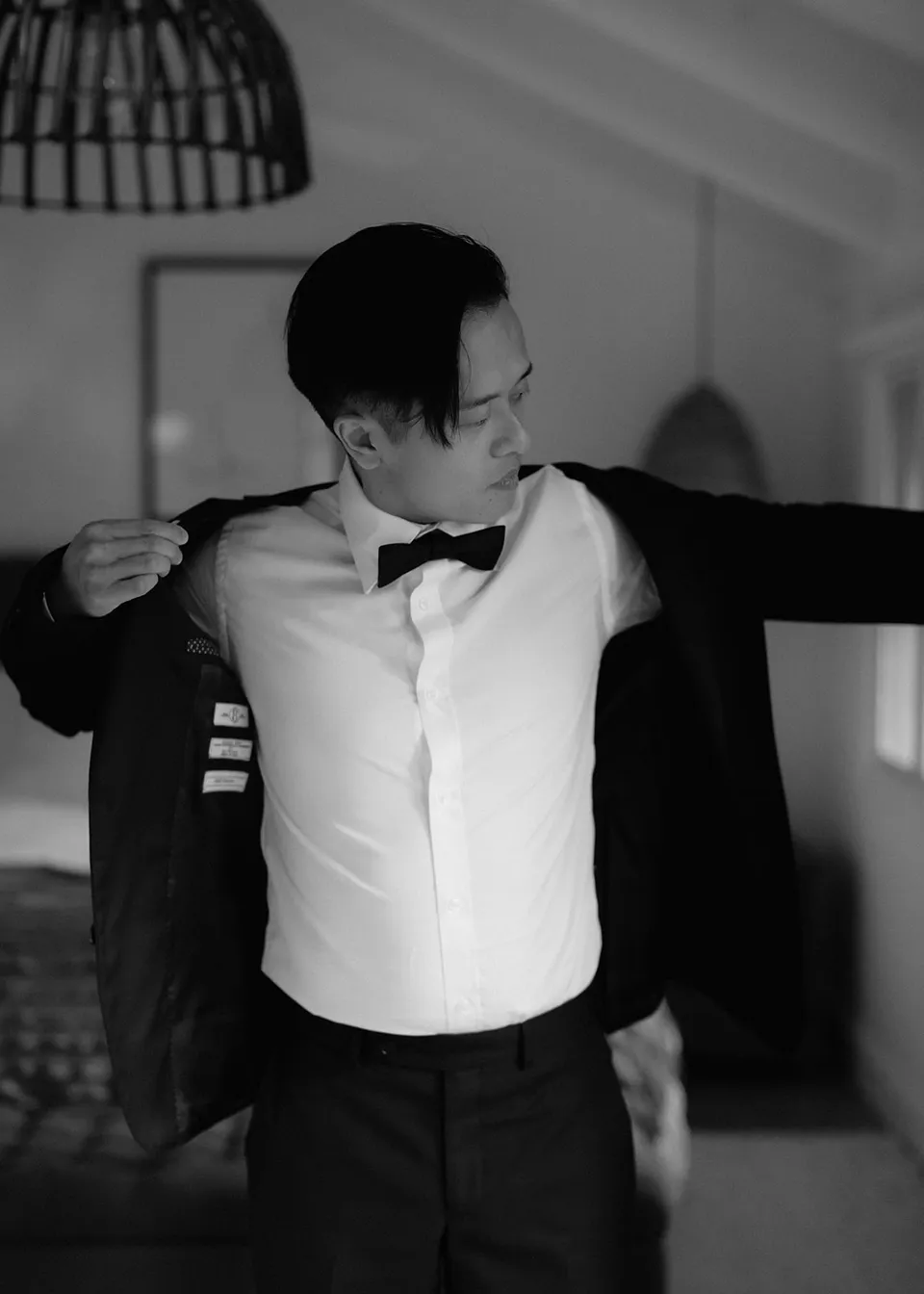 Black-and-white photograph of Jonathan putting on his jacket. At Fig Tree Villa in Ewingsdale, Byron Bay, for an Elopement Collective elopement. Photograph by Bulb Creative.