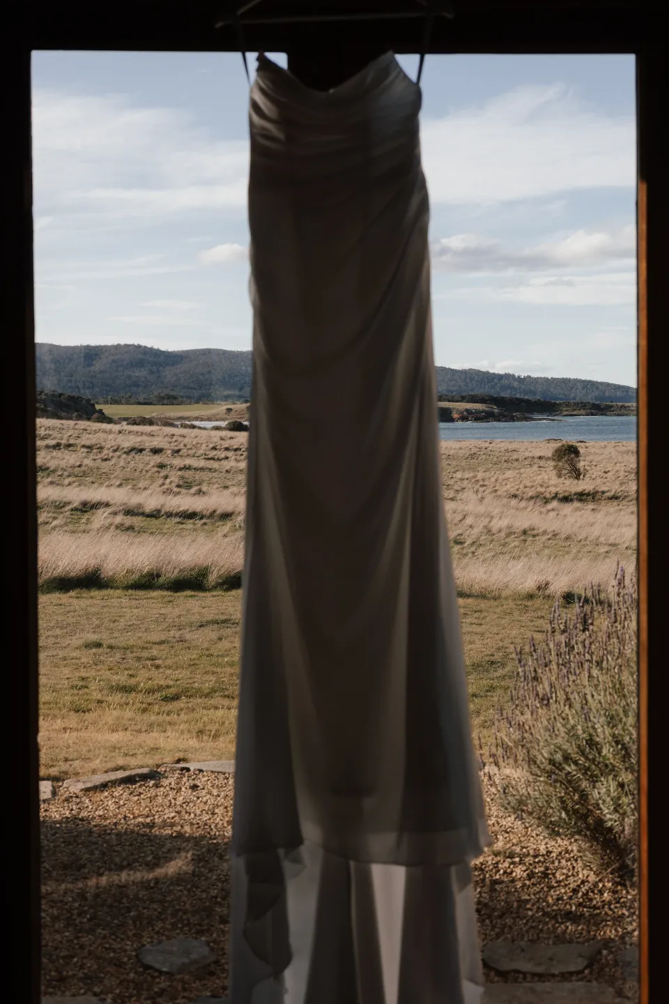 Dani’s white wedding dress hanging in a doorway overlooking the coastal grassland and bay, at Thalia Haven on Tasmania’s east coast, for an Elopement Collective elopement. Photograph by Bulb Creative.