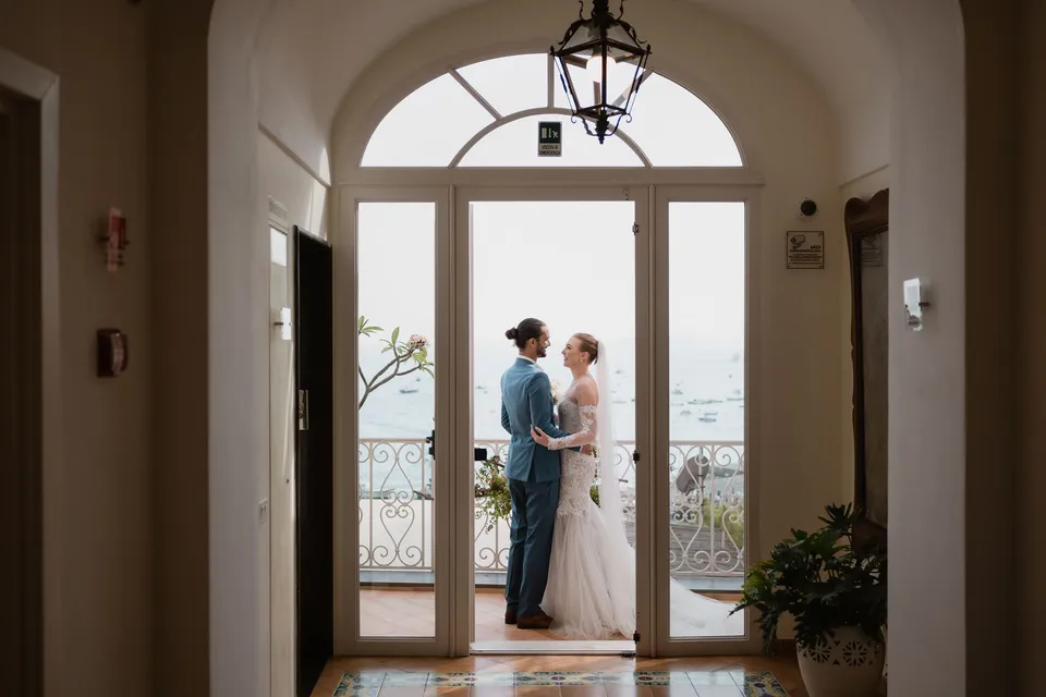 Skyler and Zach standing together in open balcony doors, framed by an arch and a hanging lantern, at Hotel Buca di Bacco in Positano, Amalfi Coast, Italy, for an Elopement Collective elopement. Photograph by Jason Corroto.