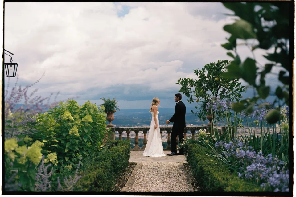 Holly and Matthew on the ceremony terrace, viewed through purple flowers and garden foliage, at Villa San Michele in Fiesole above Florence, Tuscany, Italy, for an Elopement Collective elopement. Film photograph by Khara from House of Lucie.