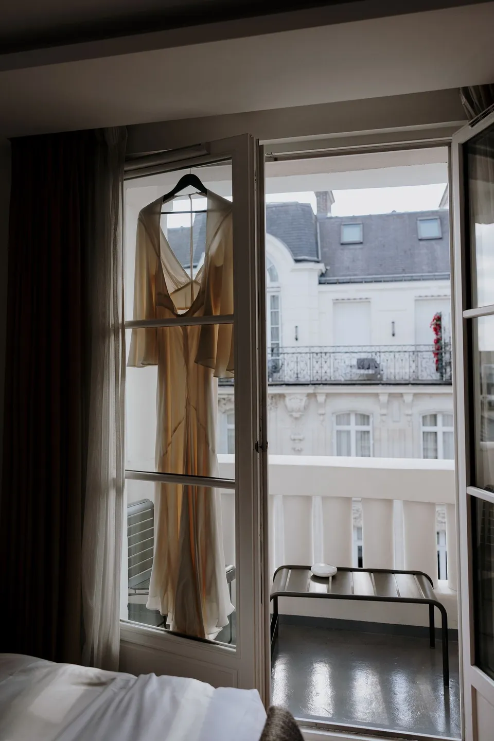 Open French doors and curtains beside a bed, with a chair near the balcony and Parisian buildings outside, at Hotel Marignan Champs-Élysées in Paris, France, during an Elopement Collective elopement. Photograph by George Bowden.
