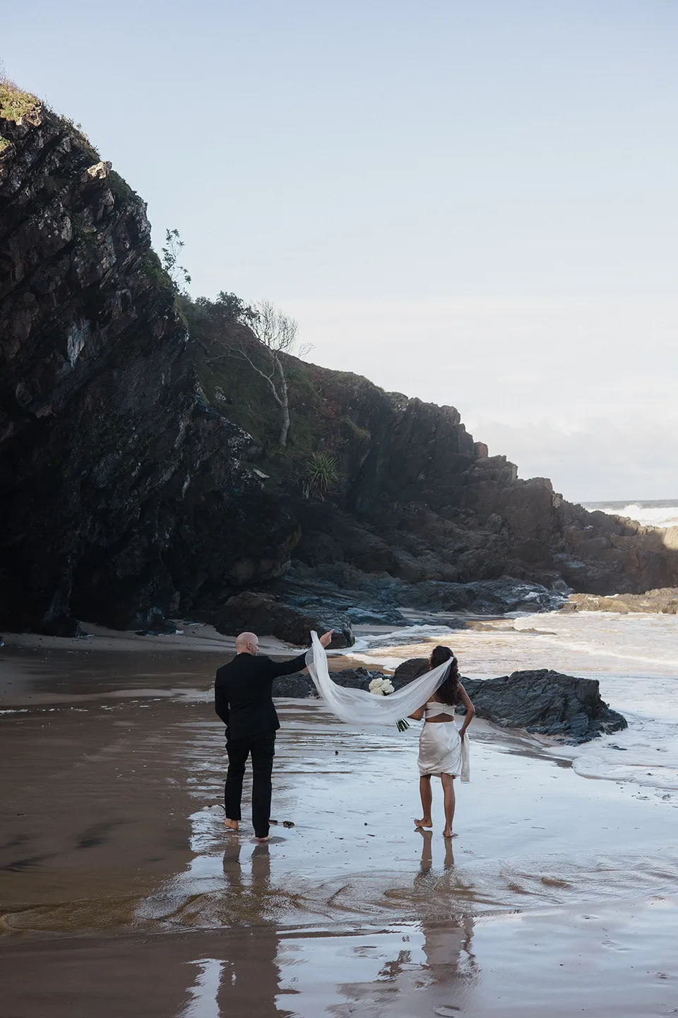 Rachel and Dane holding hands on the sand beneath the headland, with her veil lifting in the breeze. At Whites Beach, Broken Head, near Byron Bay, for an Elopement Collective elopement. Photograph by Jason Lucas.