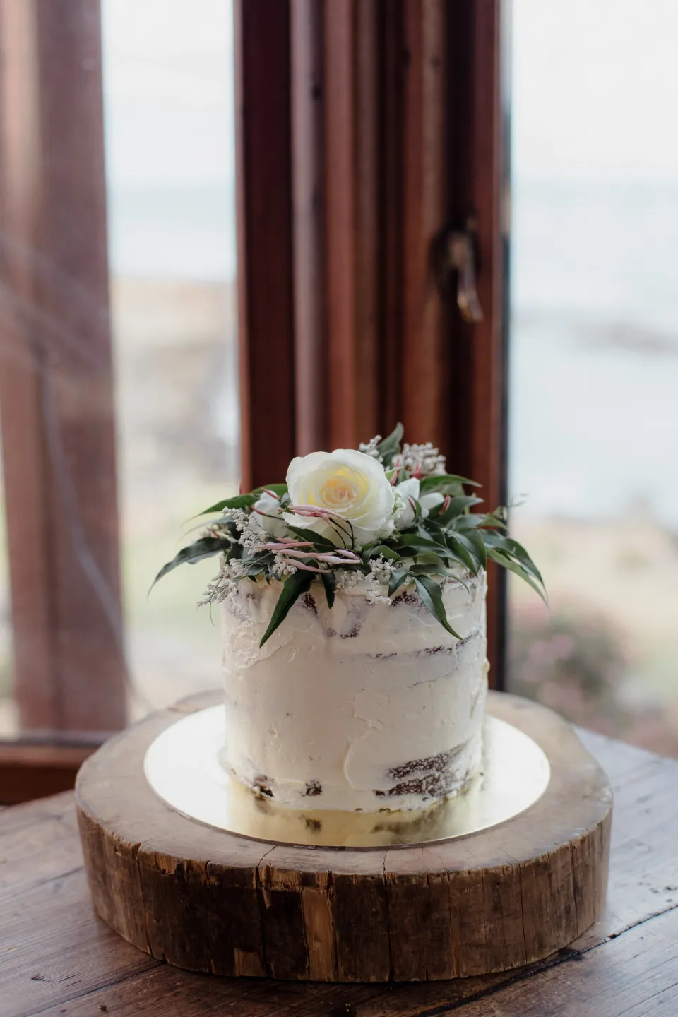 A small white wedding cake with flowers beside a timber-framed window, at Thalia Haven on the East Coast of Tasmania, Australia, for an Elopement Collective elopement. Photograph by Joey Willis.