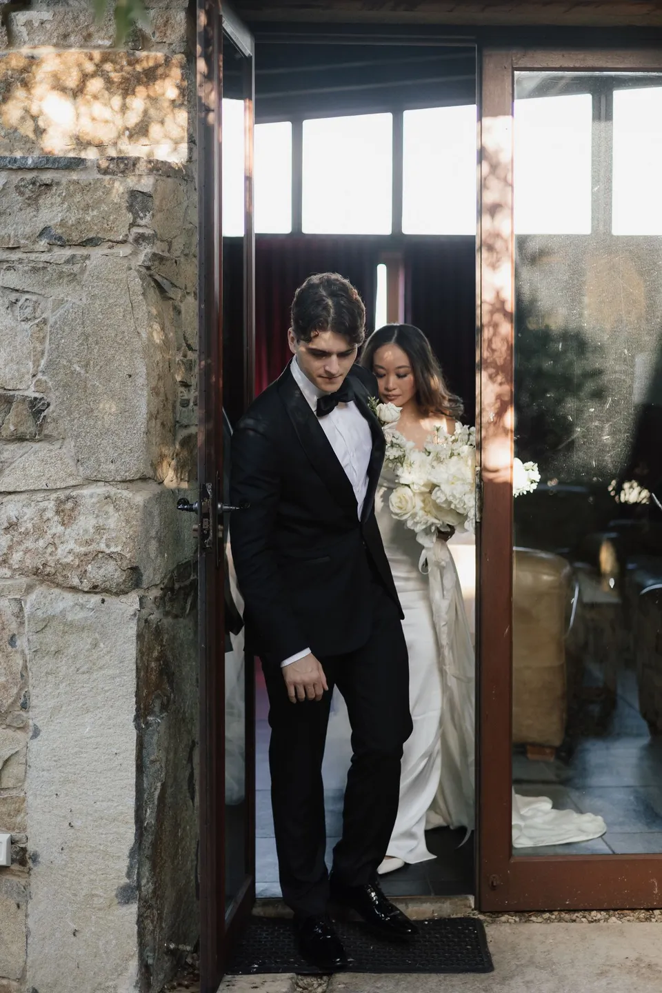 Clara and Harry standing together in the open doorway of the stone house, with her white bouquet by The Romantics, at Thalia Haven on the East Coast of Tasmania, for an Elopement Collective elopement. Photograph by Bulb Creative.