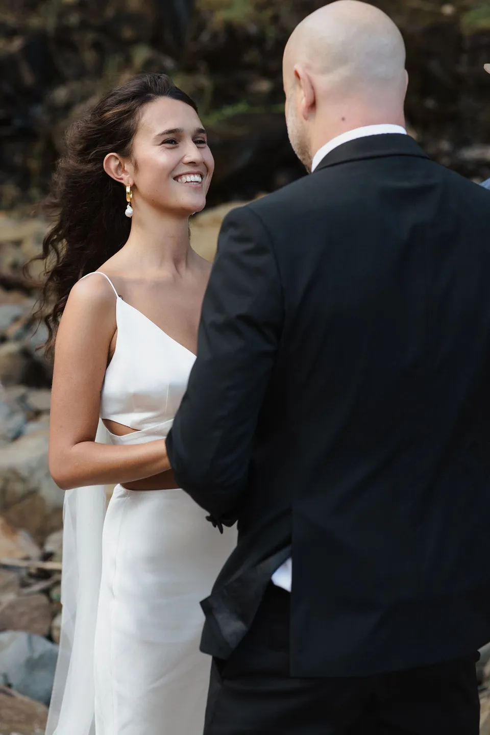 Rachel smiling at Dane with the coastal rock face behind them. At Whites Beach, Broken Head, near Byron Bay, for an Elopement Collective elopement. Photograph by Jason Lucas.