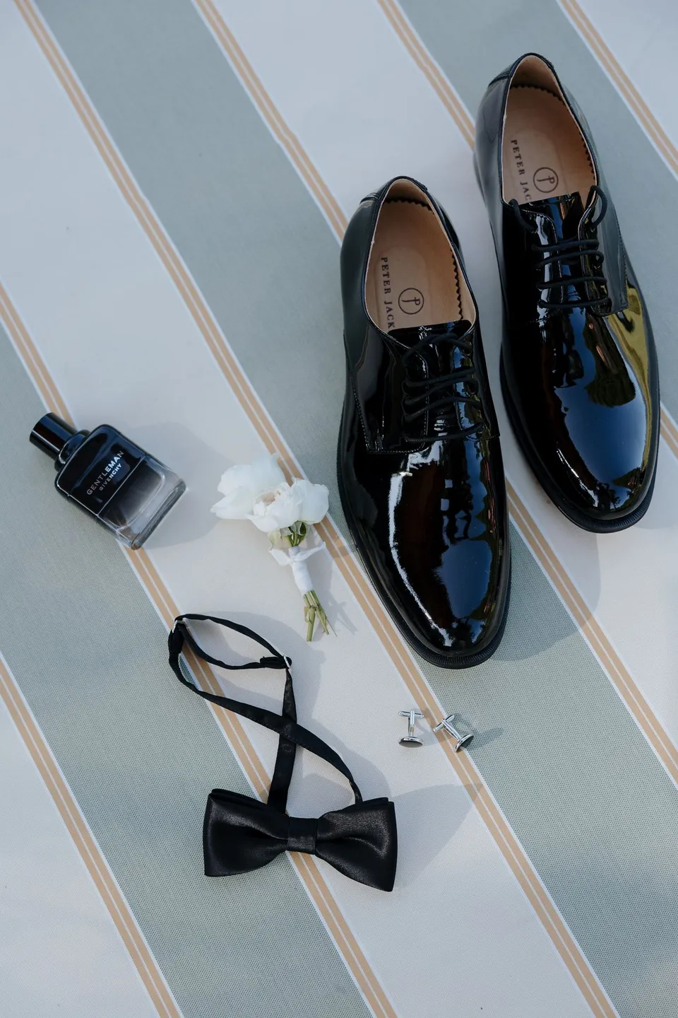 Black patent shoes, a bow tie, perfume and a white floral boutonniere by Garden of Love arranged on a striped surface, at Passalacqua in Moltrasio, Lake Como, Italy, for an Elopement Collective elopement. Photo by George Bowden.