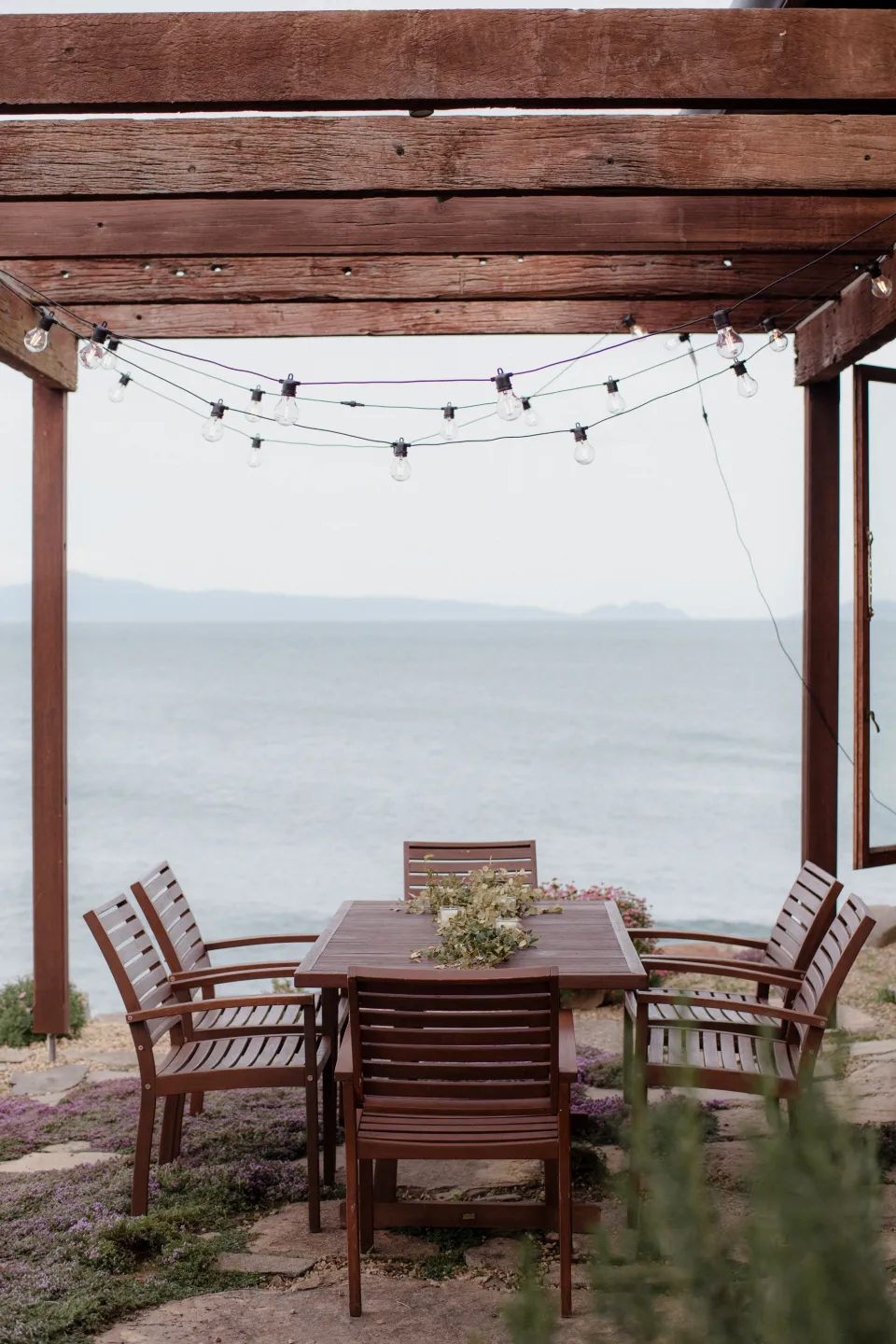 A timber dining table and chairs beneath festoon lights with the bay beyond, at Thalia Haven on the East Coast of Tasmania, Australia, for an Elopement Collective elopement. Photograph by Joey Willis.