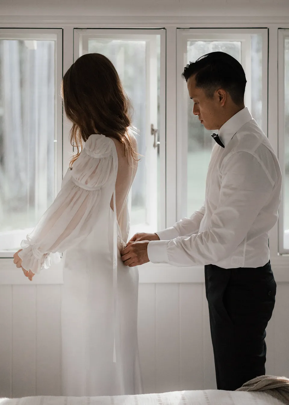 Jonathan helping fasten Elyse's wedding dress beside the window. At Fig Tree Villa in Ewingsdale, Byron Bay, for an Elopement Collective elopement. Photograph by Bulb Creative.