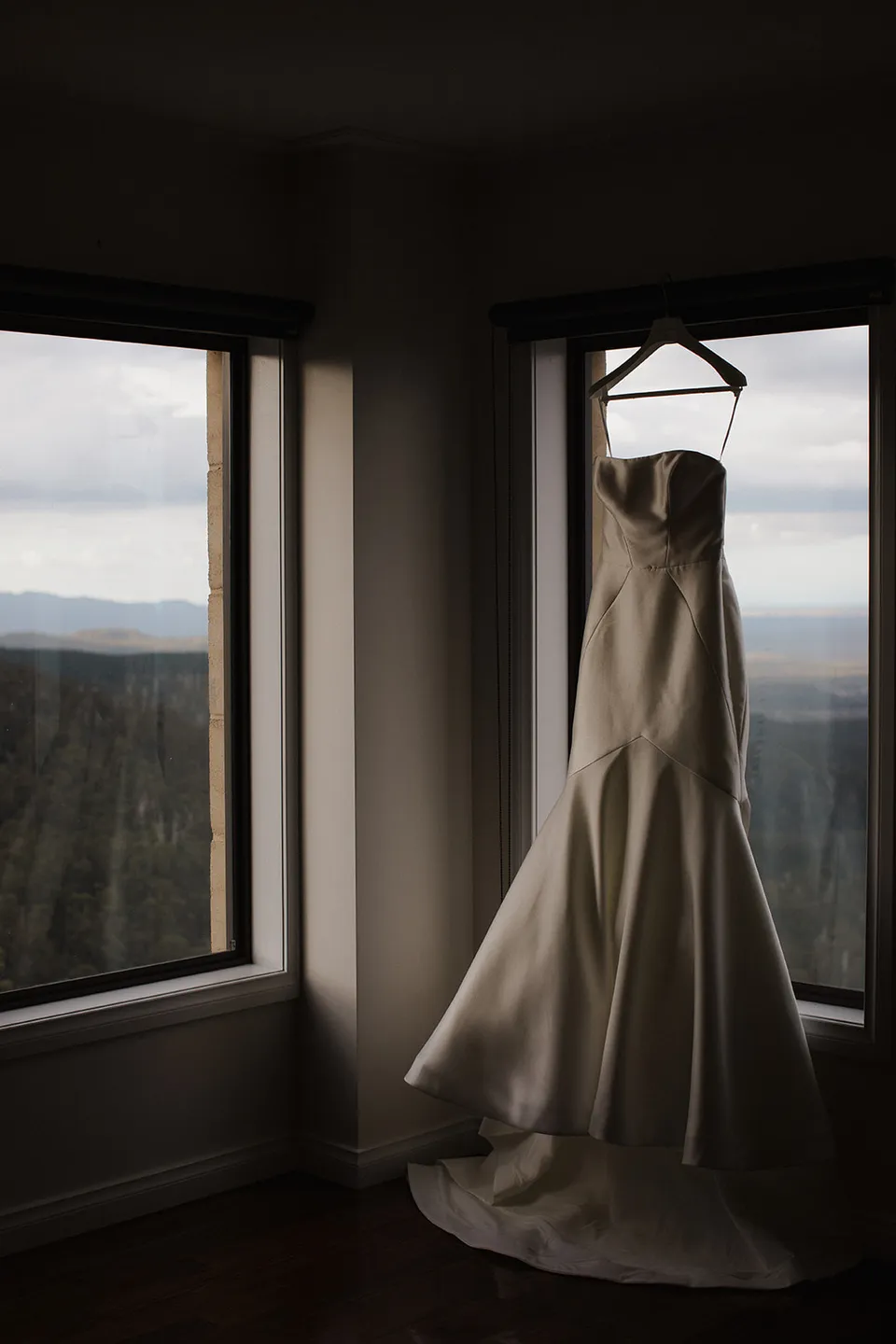 Meg’s wedding dress hanging between tall windows overlooking the forest. At The Keep on Tasmania’s east coast, for an Elopement Collective elopement. Photograph by Jenna Agius.
