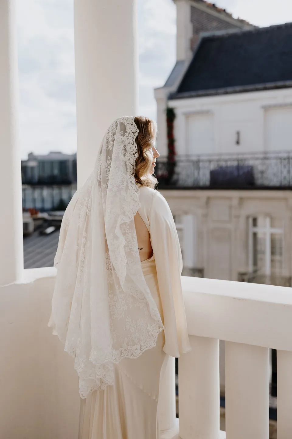 Rear view of Kara in her flowing white dress and lace veil on a stone-balustraded balcony, at Hotel Marignan Champs-Élysées in Paris, France, during an Elopement Collective elopement. Photograph by George Bowden.