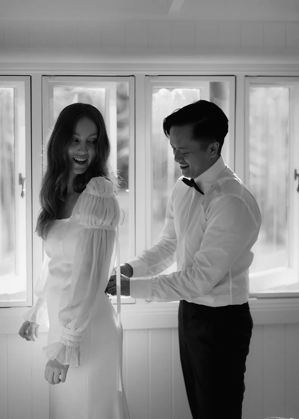 Black-and-white photograph of Elyse smiling while Jonathan helps with her dress. At Fig Tree Villa in Ewingsdale, Byron Bay, for an Elopement Collective elopement. Photograph by Bulb Creative.