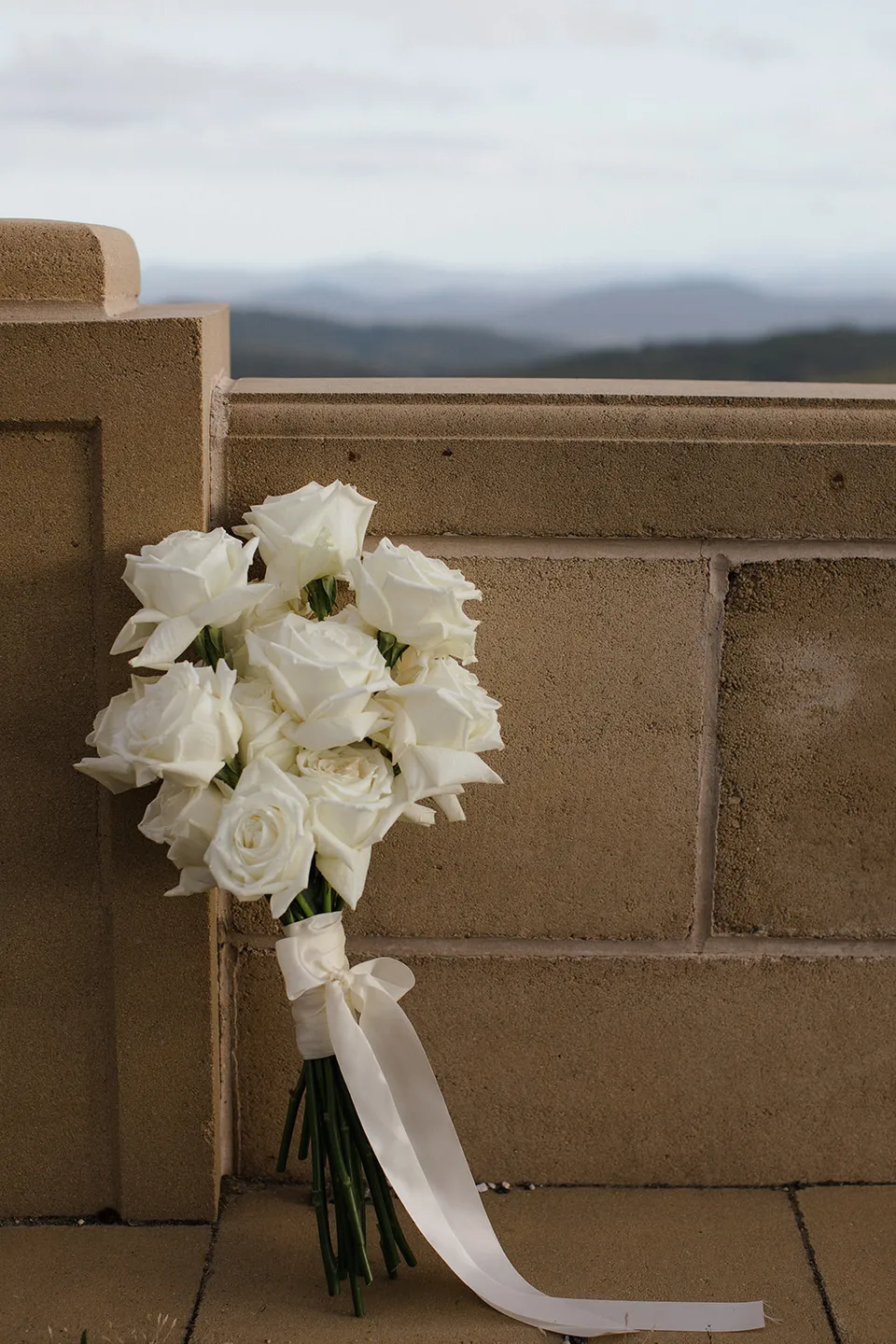 A white rose bouquet by The Romantics with long ivory ribbons resting against a sandstone wall. At The Keep on Tasmania’s east coast, for an Elopement Collective elopement. Photograph by Jenna Agius.
