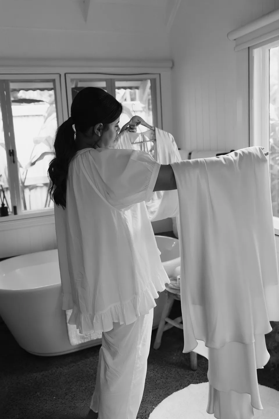 Black-and-white photograph of Laura looking at her wedding dress beside the window. At Fig Tree Villa in Ewingsdale, Byron Bay, for an Elopement Collective elopement. Photograph by George Bowden.