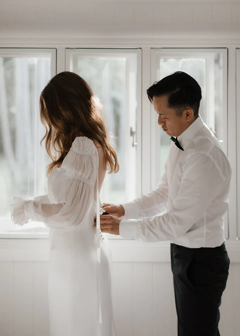 Jonathan carefully fastening the back of Elyse's dress in the window light. At Fig Tree Villa in Ewingsdale, Byron Bay, for an Elopement Collective elopement. Photograph by Bulb Creative.