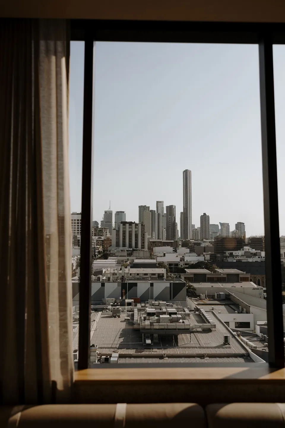 Brisbane’s skyline framed by the hotel suite window and curtains, during Danielle and Adam’s Elopement Collective elopement at The Calile Hotel in Fortitude Valley, Brisbane.