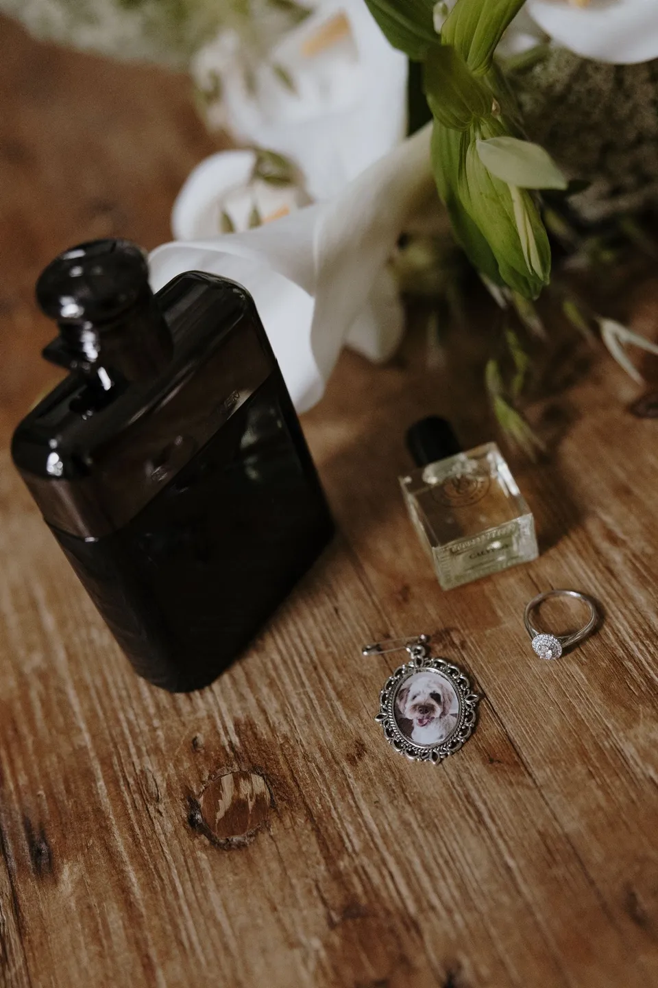 Two perfume bottles, a ring and a small framed dog portrait beside white flowers by The French Petal on a timber table. At Fig Tree Villa in Ewingsdale, Byron Bay, for an Elopement Collective elopement. Photograph by George Bowden.