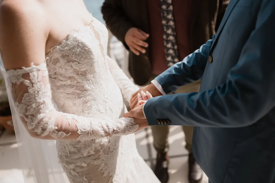 Close view of Skyler and Zach holding hands, with her lace sleeves, his blue suit and celebrant Josh Withers behind them, at Hotel Buca di Bacco in Positano, Amalfi Coast, Italy, for an Elopement Collective elopement. Photograph by Jason Corroto.