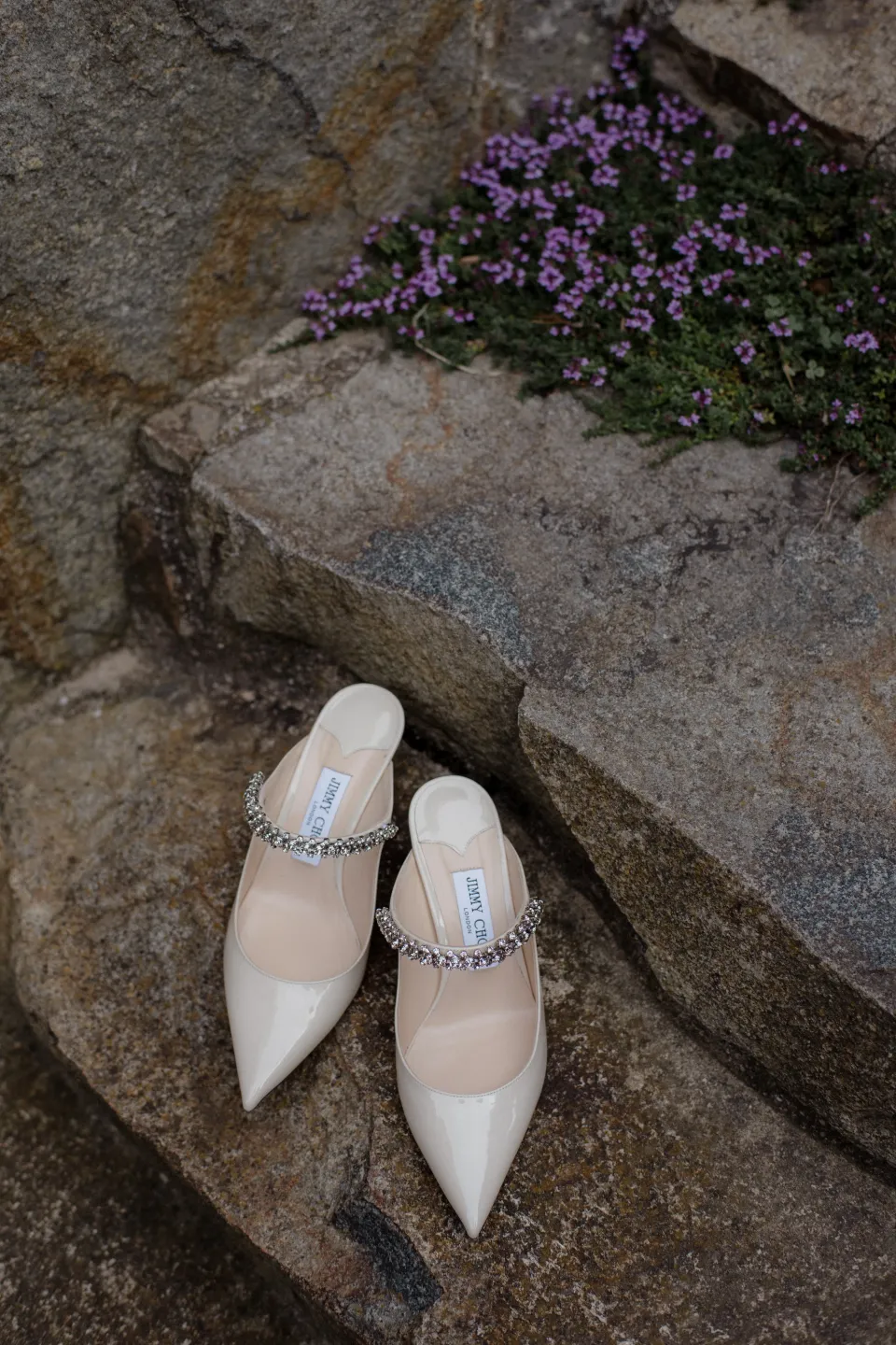 White bridal shoes resting on stone beside purple garden flowers, at Thalia Haven on the East Coast of Tasmania, Australia, for an Elopement Collective elopement. Photograph by Joey Willis.