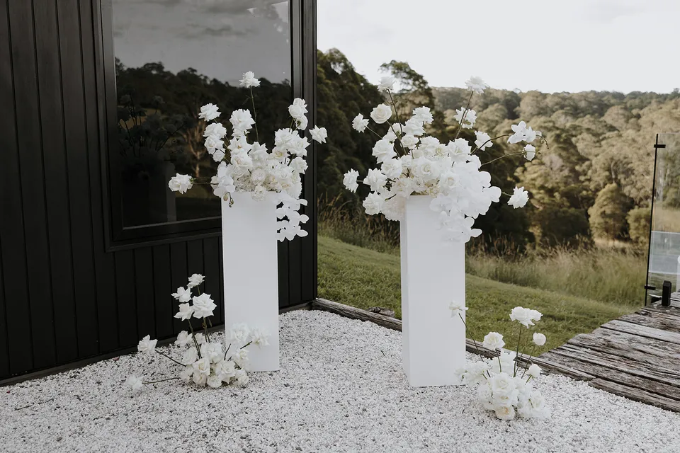 White flowers by Mrs Gibbons arranged on tall plinths and pale gravel beside the dark cabin for an Elopement Collective elopement at Roundhill Retreat in Obi Obi, Sunshine Coast Hinterland.