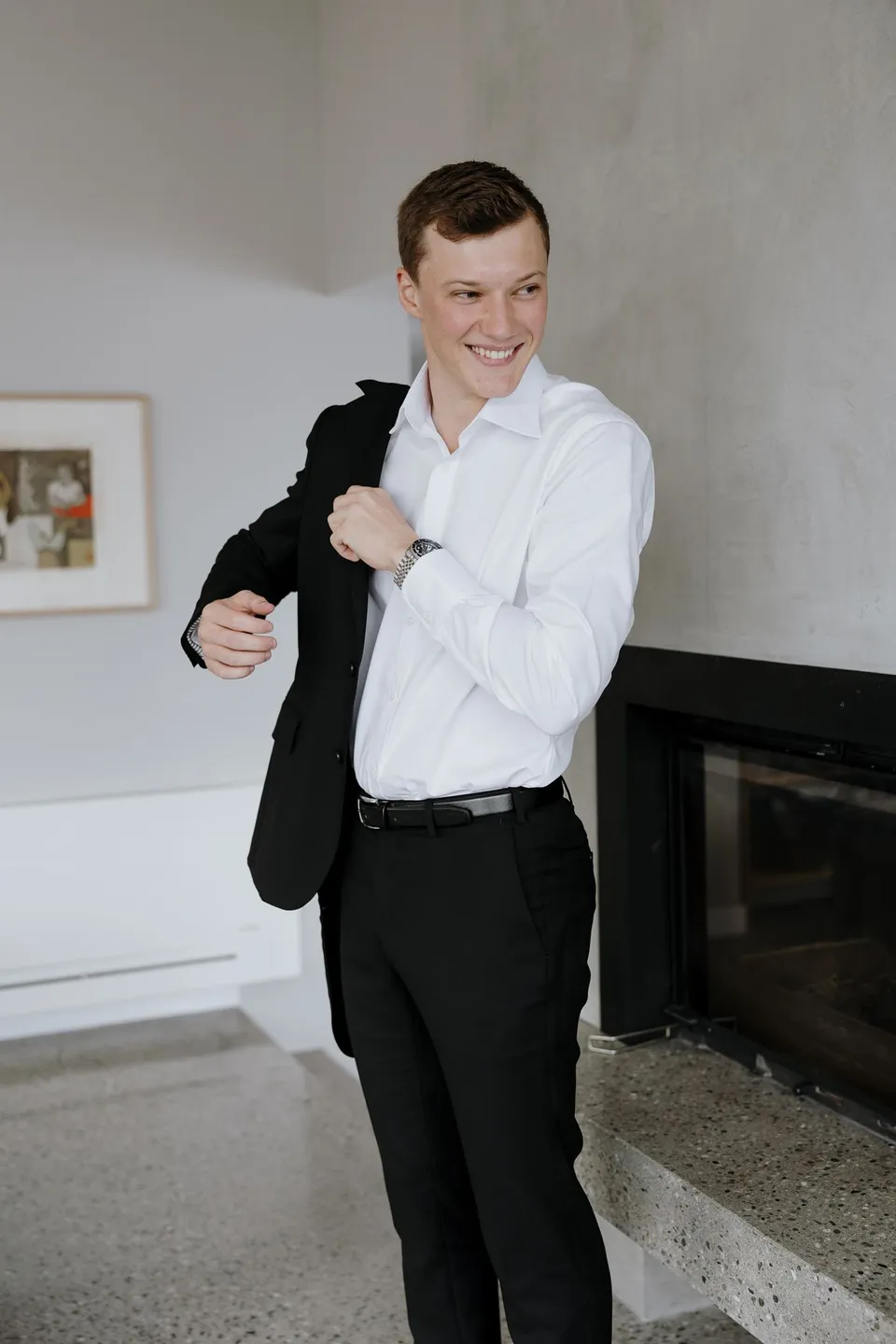 Hamish smiling as he puts on his black suit jacket over a white shirt, at their Airbnb in Fernhill, Queenstown, New Zealand, for an Elopement Collective Queenstown elopement. Photo by George Bowden.
