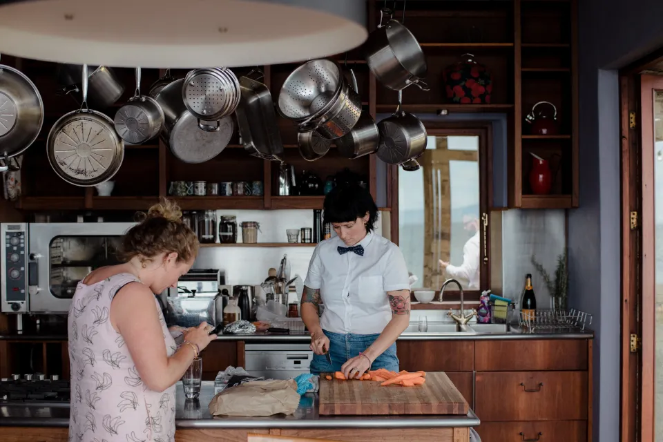 Two people preparing food in the kitchen beneath hanging cookware, at Thalia Haven on the East Coast of Tasmania, Australia, for an Elopement Collective elopement. Photograph by Joey Willis.