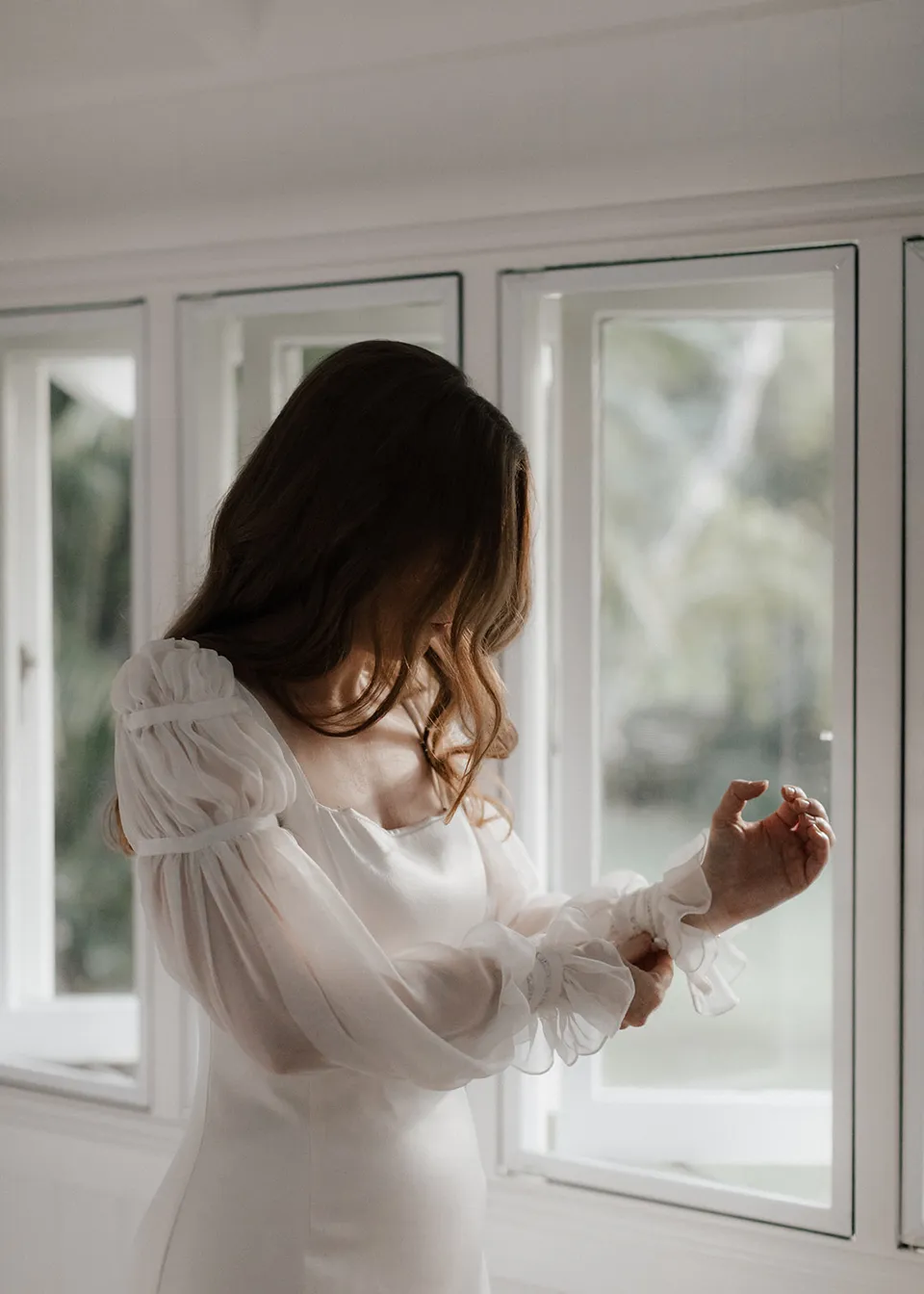 Elyse looking down while arranging her long dress sleeve beside the window. At Fig Tree Villa in Ewingsdale, Byron Bay, for an Elopement Collective elopement. Photograph by Bulb Creative.