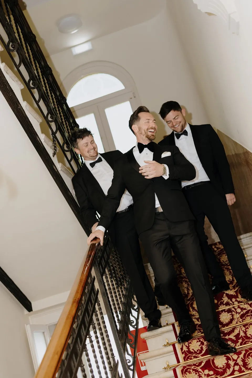 Three men in black tuxedos descending an ornate staircase with a patterned red carpet at Hotel Villa Marie in Tremezzo, at Lake Como in Lombardy, Italy, for an Elopement Collective elopement. Photograph by George Bowden.